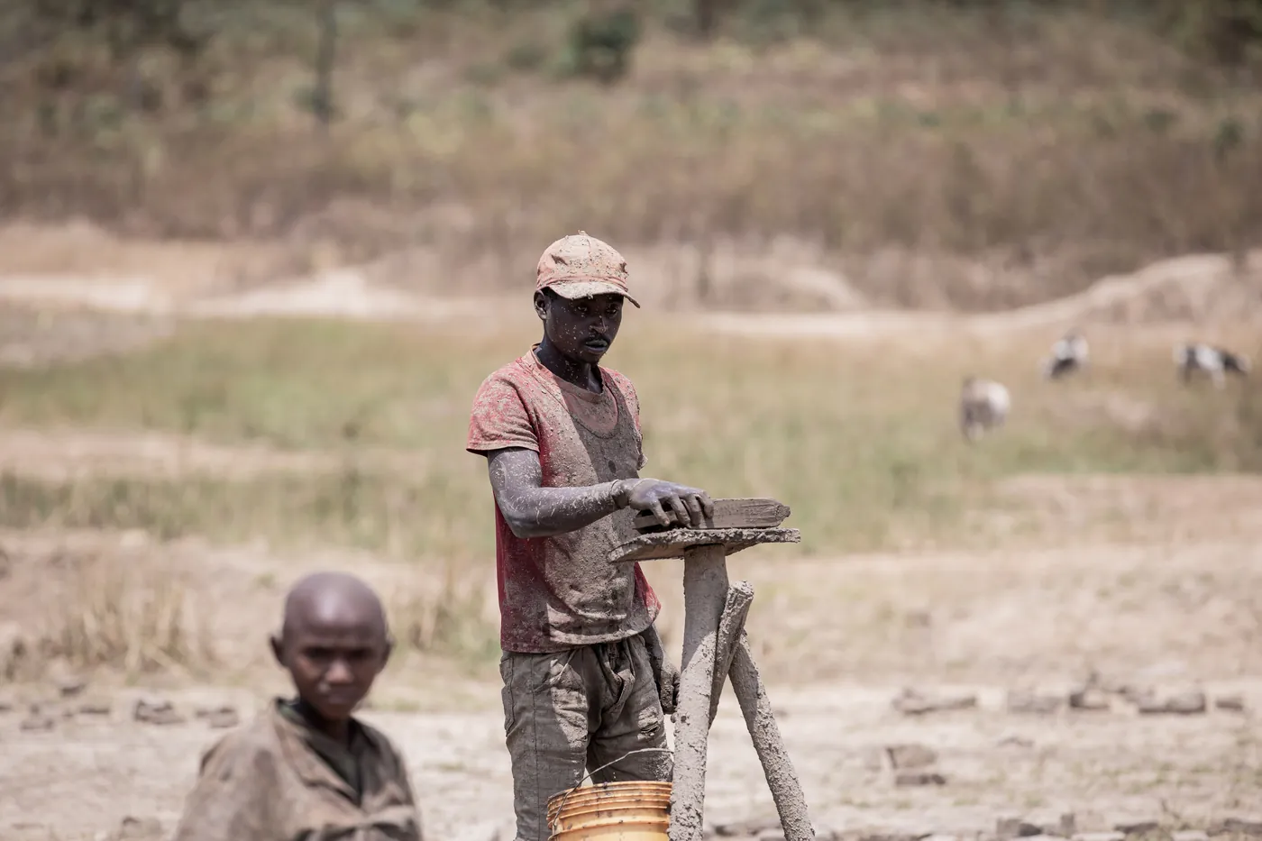 A person in a cap standing beside a wooden support, with another person in the foreground. a-person-in-a-cap-standing-beside-a-wooden-support,-with-another-person-in-the-foreground-for-owaid-orphan-and-widow-support