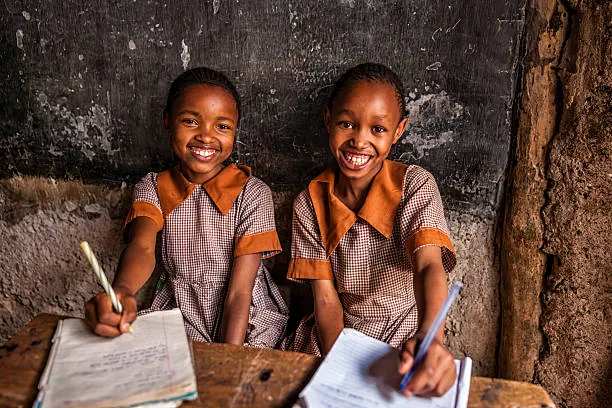Two students in matching uniforms smiling at a wooden classroom desk. two-students-in-matching-uniforms-smiling-at-a-wooden-classroom-desk-for-owaid-orphan-and-widow-support