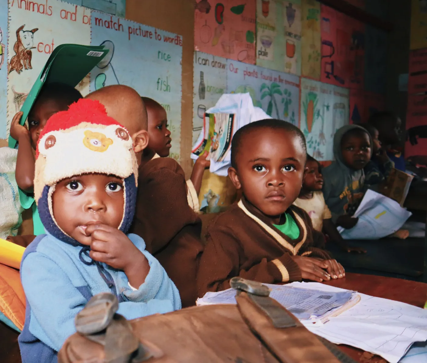 Young children sitting at a classroom table with notebooks and learning posters behind them. young-children-sitting-at-a-classroom-table-with-notebooks-and-learning-posters-behind-them-for-owaid-orphan-and-widow-support