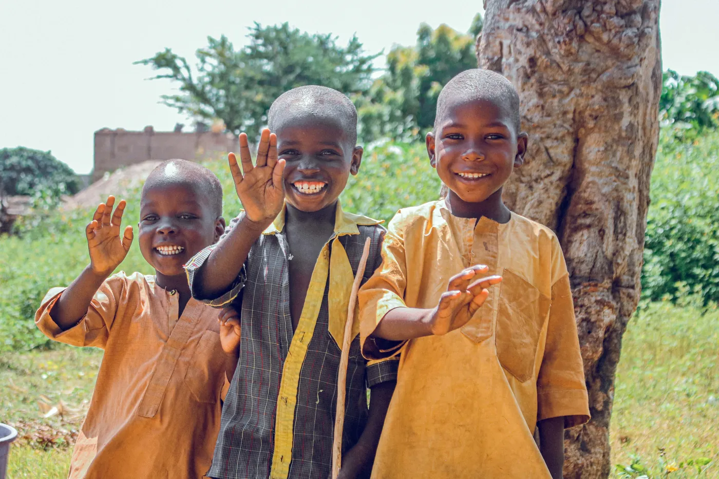 Three smiling children waving outdoors beside a tree. three-smiling-children-waving-outdoors-beside-a-tree-for-owaid-orphan-and-widow-support