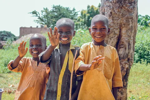three-smiling-children-waving-outdoors-beside-a-tree
