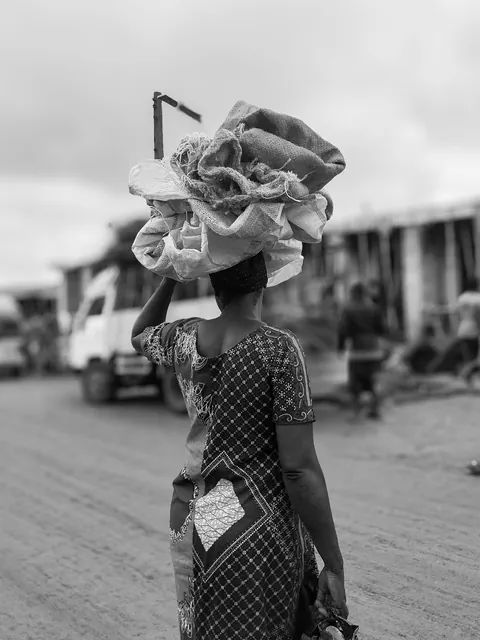 a-black-and-white-photograph-of-a-woman-carrying-a-large-bundle-of-goods-on-her-head