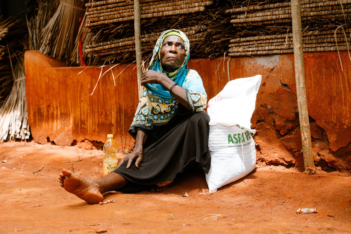 an-older-woman-seated-beside-a-white-sack-outside-a-woven-and-earthen-building-for-owaid-orphan-and-widow-support